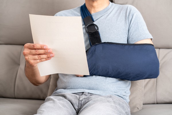 A man sitting on a couch with his arm in a blue medical sling, holding and reviewing a document from the insurance company.