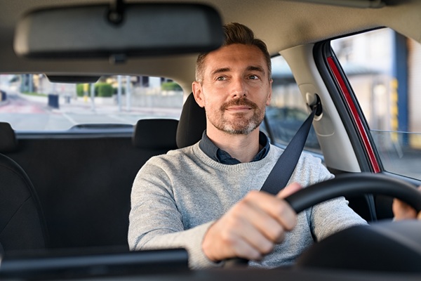 A man with light brown hair and a beard wearing a grey sweater and a seatbelt while calmly driving a car.