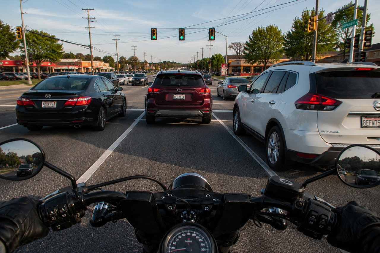 A first-person view from a motorcycle stopped at a busy multi-lane intersection during late afternoon, showing the rider positioned directly behind several cars and SUVs. This perspective highlights the common traffic congestion and vehicle proximity that contribute to motorcycle crash risks and potential injuries in Maryland.