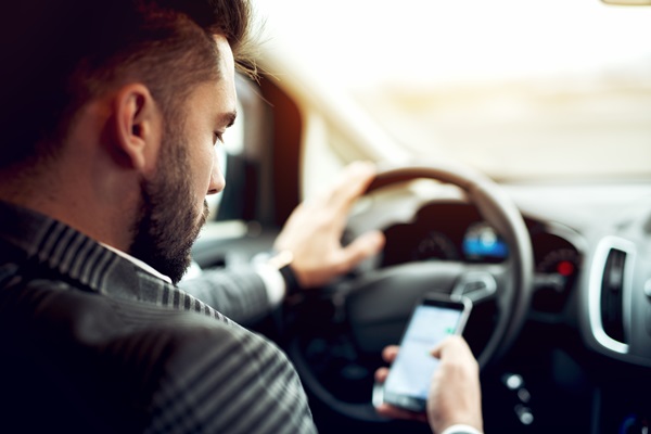A man with a beard and dark hair texting on his smartphone while driving a car, looking down at the screen instead of the road.
